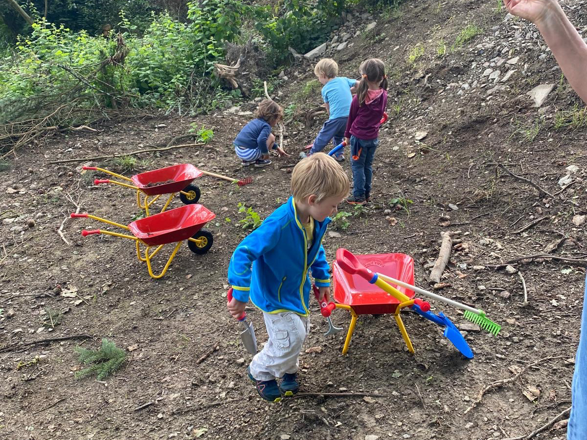 Mehrere Kinder spielen im Freien mit bunten Kinderschubkarren, Schaufeln und Rechen auf einem naturbelassenen Boden – sie graben, sammeln und entdecken gemeinsam.