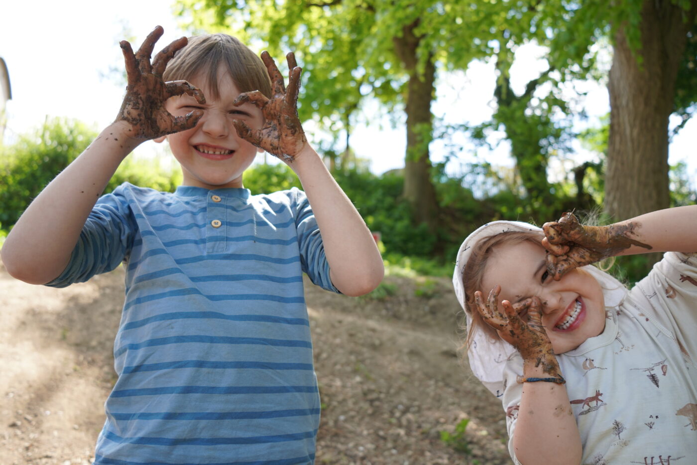 Zwei lachende Kinder zeigen mit schlammverschmierten Händen eine Brillen-Geste vor den Augen – voller Freude beim Spielen im Wald.