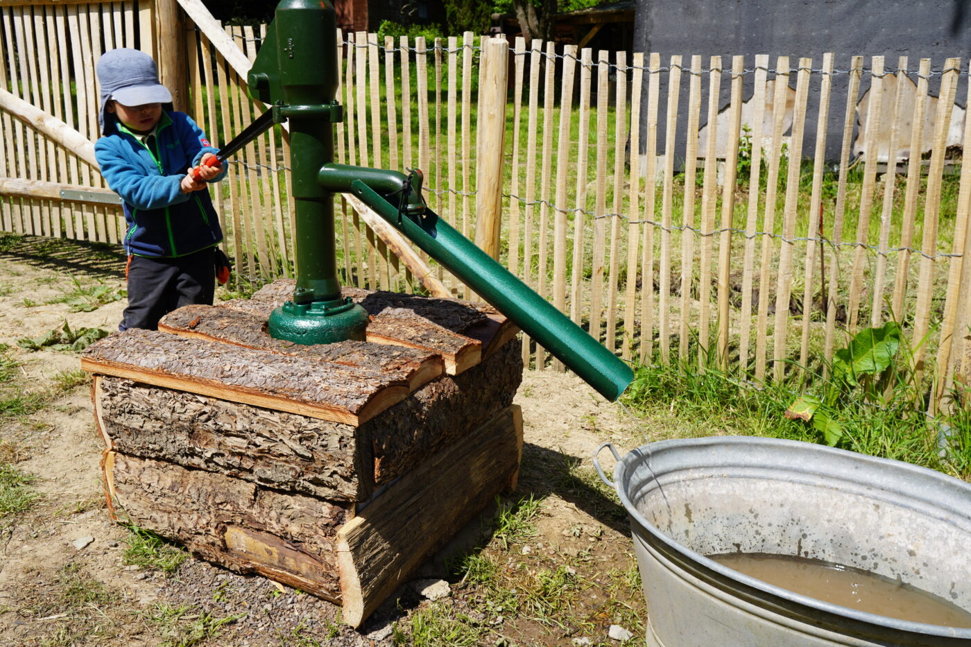 Ein Kind pumpt Wasser mit einer grünen Handpumpe, die auf einem Sockel aus Baumstämmen montiert ist. Das Wasser läuft in eine große, verzinkte Metallwanne. Im Hintergrund ist ein Holzzaun und ein naturbelassener Gartenbereich zu sehen.