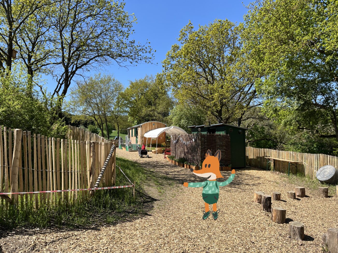 Eingangsbereich der Waldkita Mayen mit Holzhütten, Sonnenschirm und Naturzaun bei sonnigem Wetter, ergänzt durch eine gezeichnete Fuchsfigur mit Pullover in der Bildmitte.
