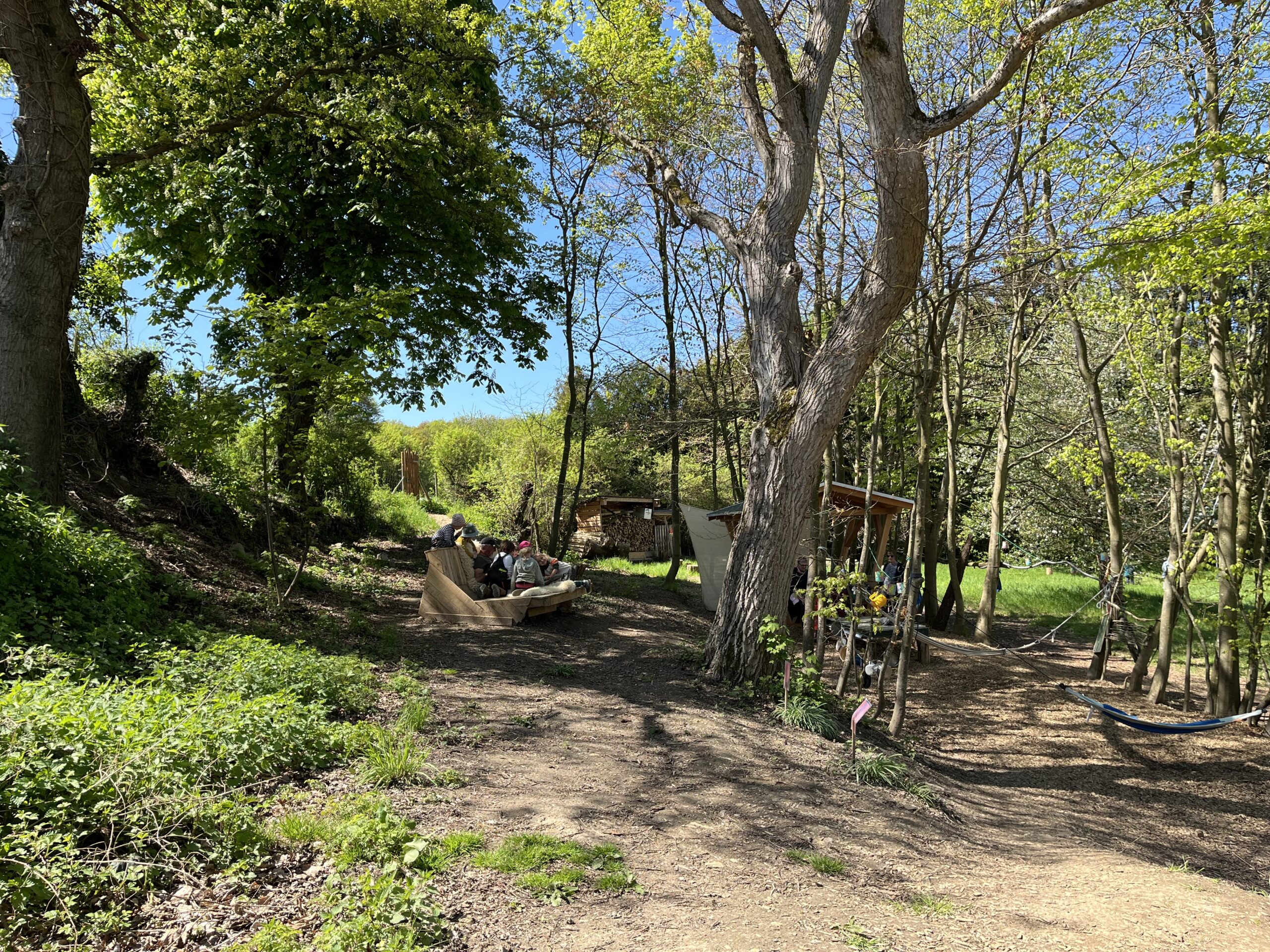 Ein sonniger Waldplatz mit Hängematte, Unterstand und Sitzbank, auf der mehrere Kinder entspannt beisammensitzen – naturnaher Alltag im Waldkindergarten.